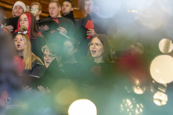 The photo shows a choir of people singing. In the foreground, blurred lights from Christmas tree lights and fragments of branches can be seen.